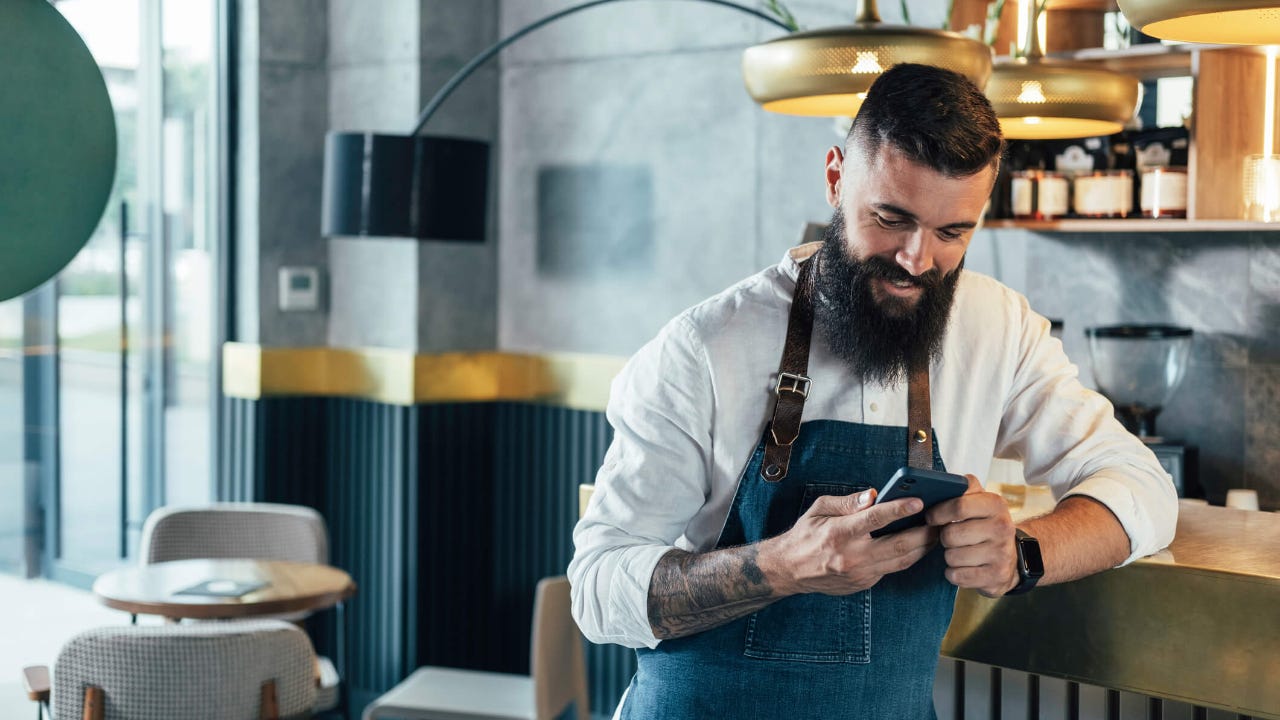 Young adult male wearing an apron at a cafe smiles while using his smartphone