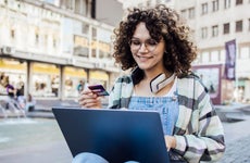 Young adult woman wearing headphones holds her credit card while using her laptop on a city street