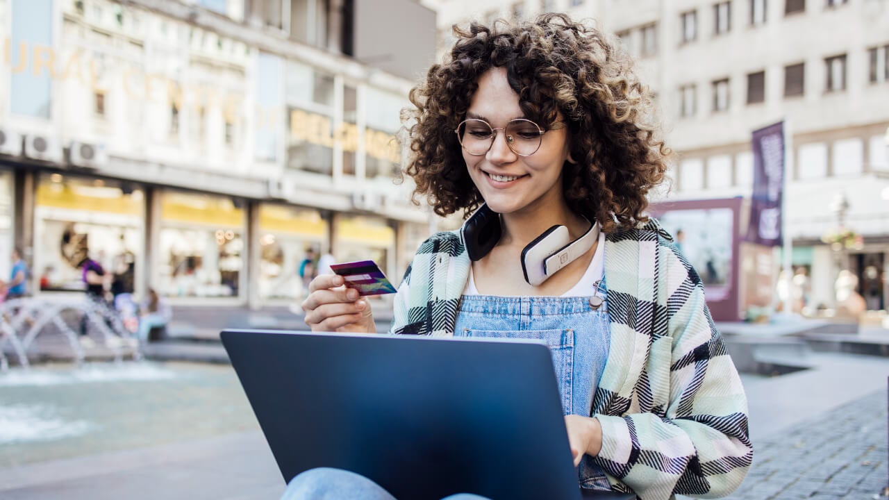 Young adult woman wearing headphones holds her credit card while using her laptop on a city street