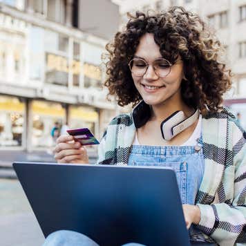 Young adult woman wearing headphones holds her credit card while using her laptop on a city street