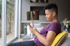 Adult woman sits on the couch in front of her living room window as she uses her smartphone