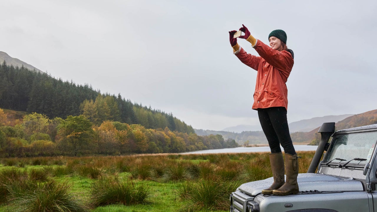 Young traveler standing on her car hood takes a photo of the forest landscape around her
