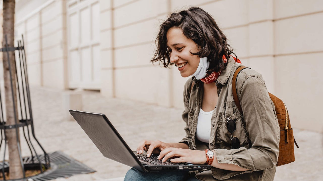 Young woman wearing face mask sits on a city curb and smiles as she uses her laptop