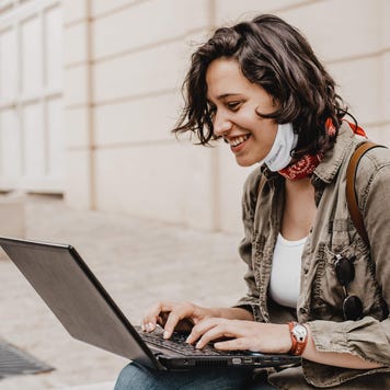 Young woman wearing face mask sits on a city curb and smiles as she uses her laptop