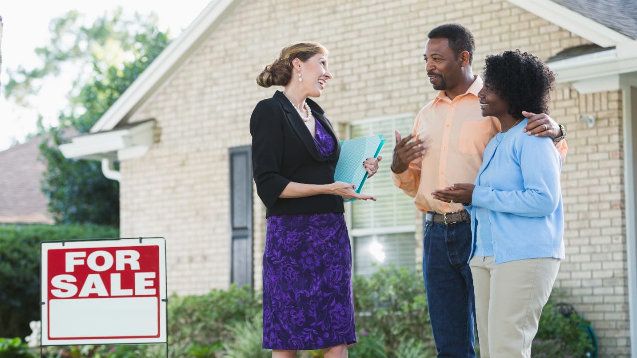 A real estate agent standing in front of a house with a FOR SALE sign in the yard, talking with an African American couple who could be the homeowers, or potential buyers. The agent is wearing a black jacket and purple dress, carrying a blue binder.
