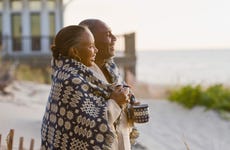Senior couple with a blanket wrapped around them laugh as they look out at the ocean from a beach house