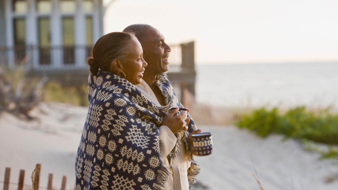 Senior couple with a blanket wrapped around them laugh as they look out at the ocean from a beach house