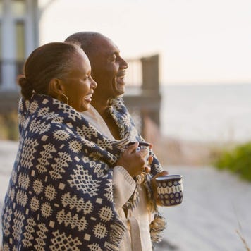 Senior couple with a blanket wrapped around them laugh as they look out at the ocean from a beach house