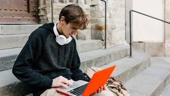 Young adult man with headphones and a backpack uses his laptop while sitting outside