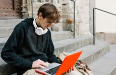 Young adult man with headphones and a backpack uses his laptop while sitting outside