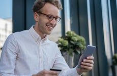Young smiling businessman holding credit card, using mobile app for online shopping. Happy handsome freelancer check card balance, receive payment. Successful business concept