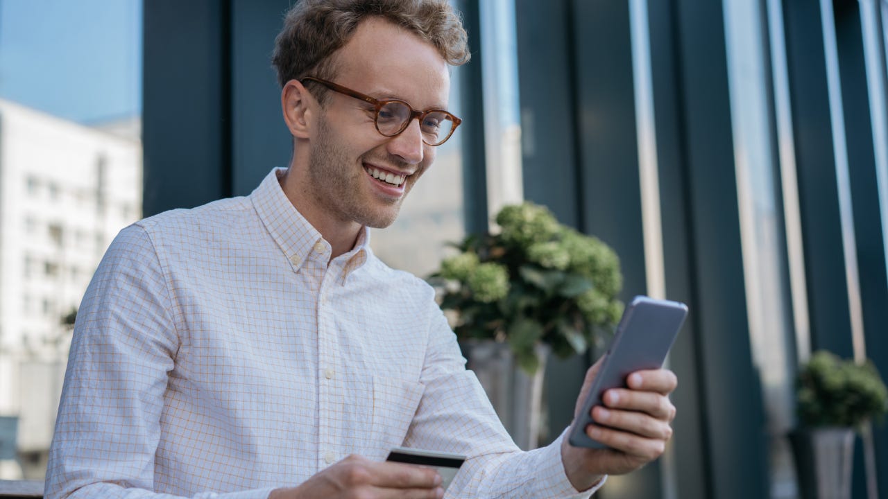 Young smiling businessman holding credit card, using mobile app for online shopping. Happy handsome freelancer check card balance, receive payment. Successful business concept