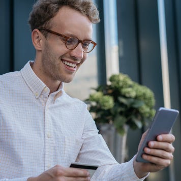 Young smiling businessman holding credit card, using mobile app for online shopping. Happy handsome freelancer check card balance, receive payment. Successful business concept