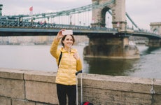Tourist woman with suitcase takes selfie in front of a bridge in Budapest