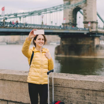 Tourist woman with suitcase takes selfie in front of a bridge in Budapest