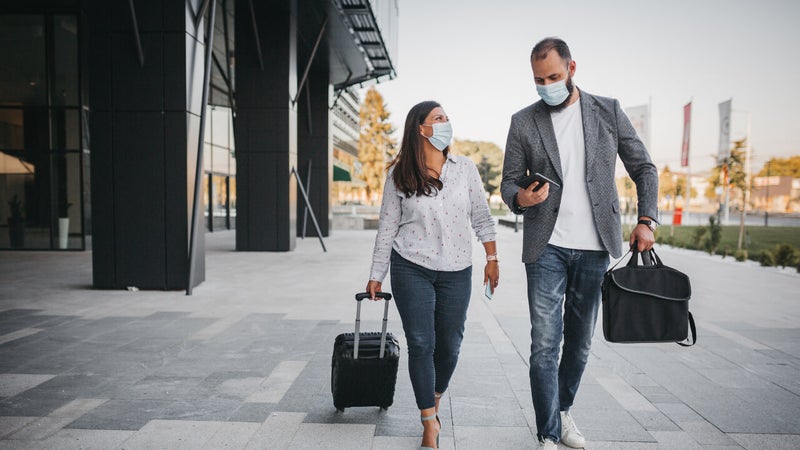 An adult woman and adult man both dressed in business casual attire and masks walk together with luggage outside of an airport terminal