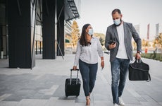 An adult woman and adult man both dressed in business casual attire and masks walk together with luggage outside of an airport terminal