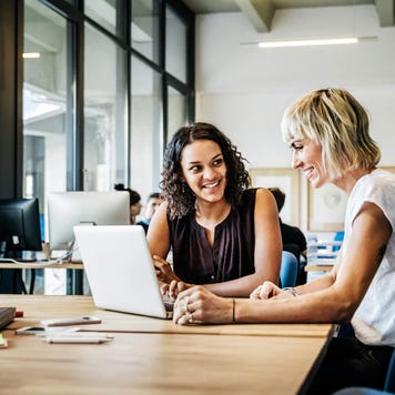 Two casual businesswomen sit at a conference table and view a laptop screen together while smiling