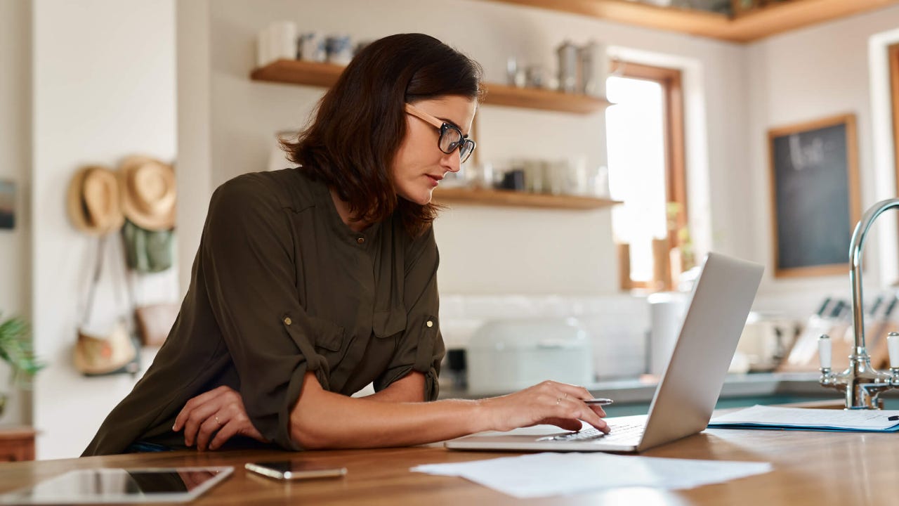 Adult woman with glasses focuses intently on her laptop screen in a kitchen