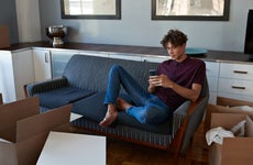 Young man scrolling on smartphone in apartment full of boxes