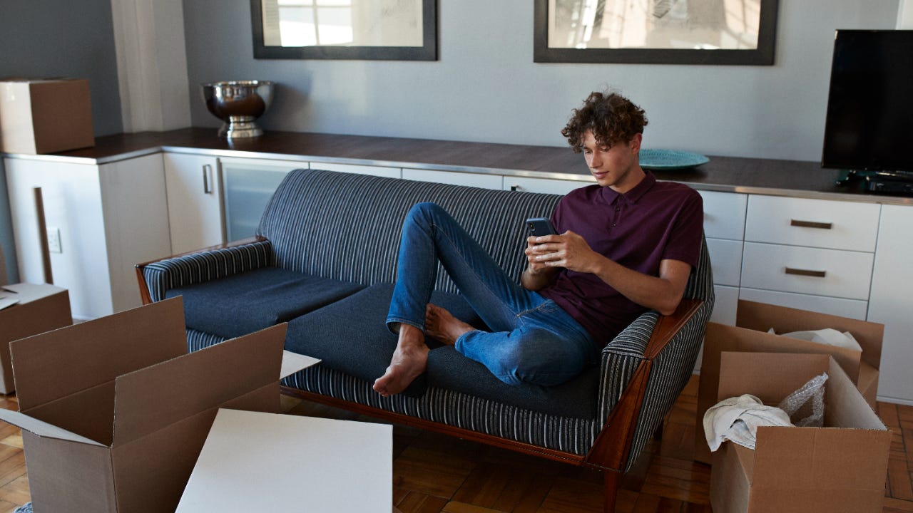 Young man scrolling on smartphone in apartment full of boxes