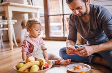 Mature father with small toddler daughter sitting on the floor indoors, eating fruit.