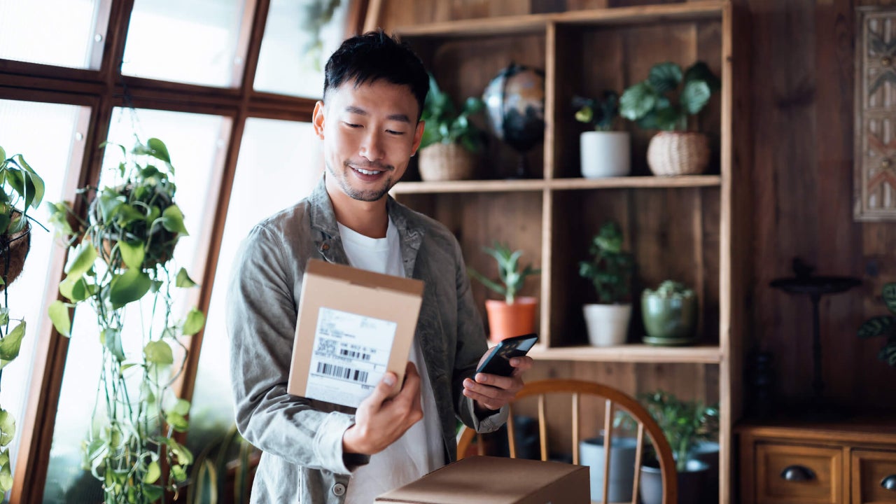 Smiling young Asian man with smartphone, receiving delivered packages from online purchases at home