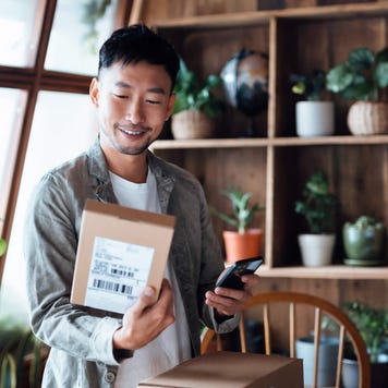 Smiling young Asian man with smartphone, receiving delivered packages from online purchases at home