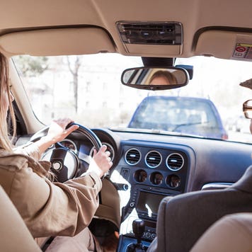 View from the backseat of a car as an older couple sits in the driver and passenger seats. The road ahead can be seen through the windshield and there is a car in front of them.