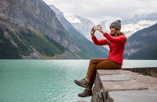A woman taking a photo on her cell phone while sitting on a rocky ledge overlooking a lake