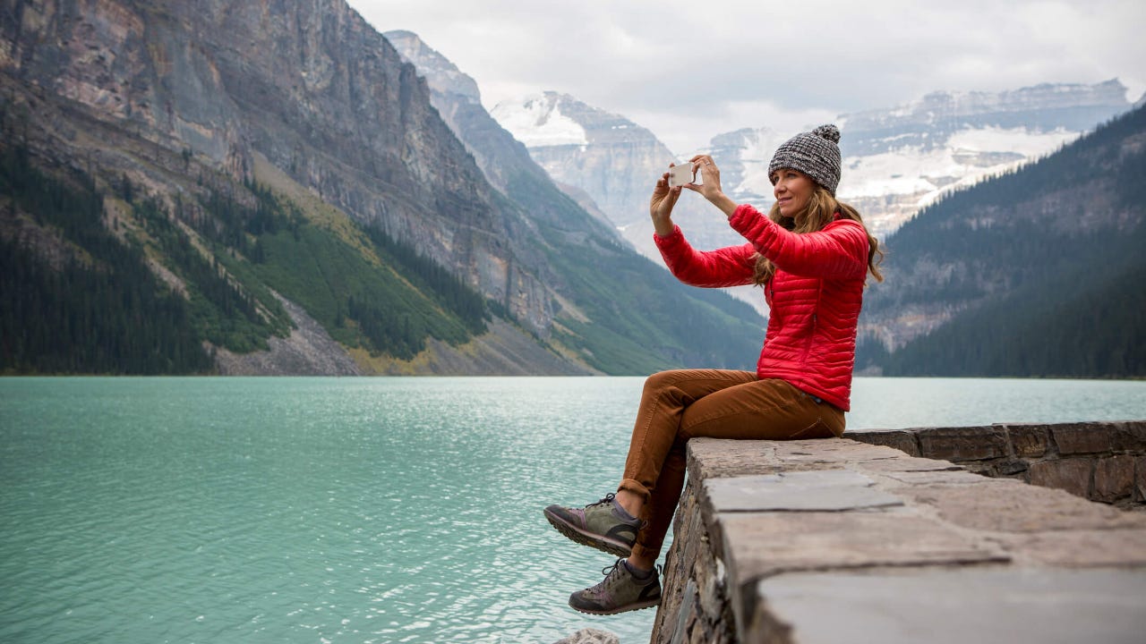 A woman taking a photo on her cell phone while sitting on a rocky ledge overlooking a lake