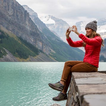 A woman taking a photo on her cell phone while sitting on a rocky ledge overlooking a lake