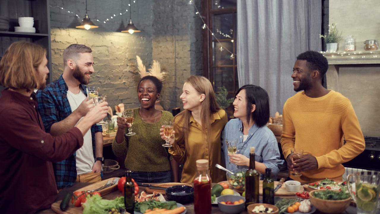 Multi-ethnic group of smiling young people enjoying dinner together standing at table in modern interior and holding wine glasses