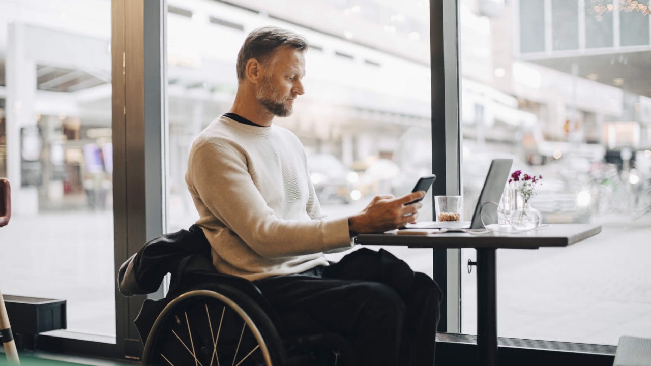 Mature businessman in wheelchair using smart phone while sitting with laptop at table in restaurant