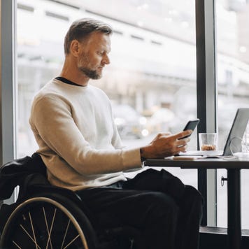 Mature businessman in wheelchair using smart phone while sitting with laptop at table in restaurant