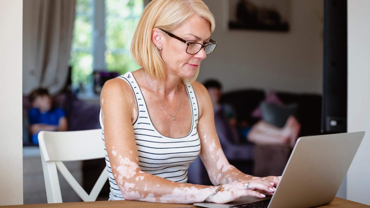 Blonde woman sitting her dining table at home with a laptop open in front of her