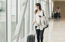 Young adult woman walks down concourse with suitcase, mask and phone