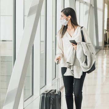 Young adult woman walks down concourse with suitcase, mask and phone