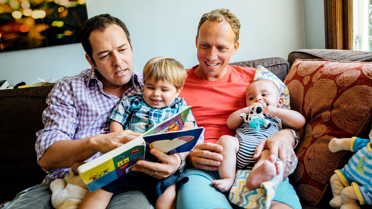 male couple reading a book with their sons on the sofa at home