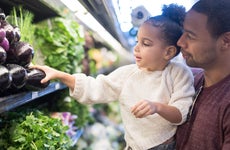 Father holds young daughter up to pick out eggplant at a grocery store