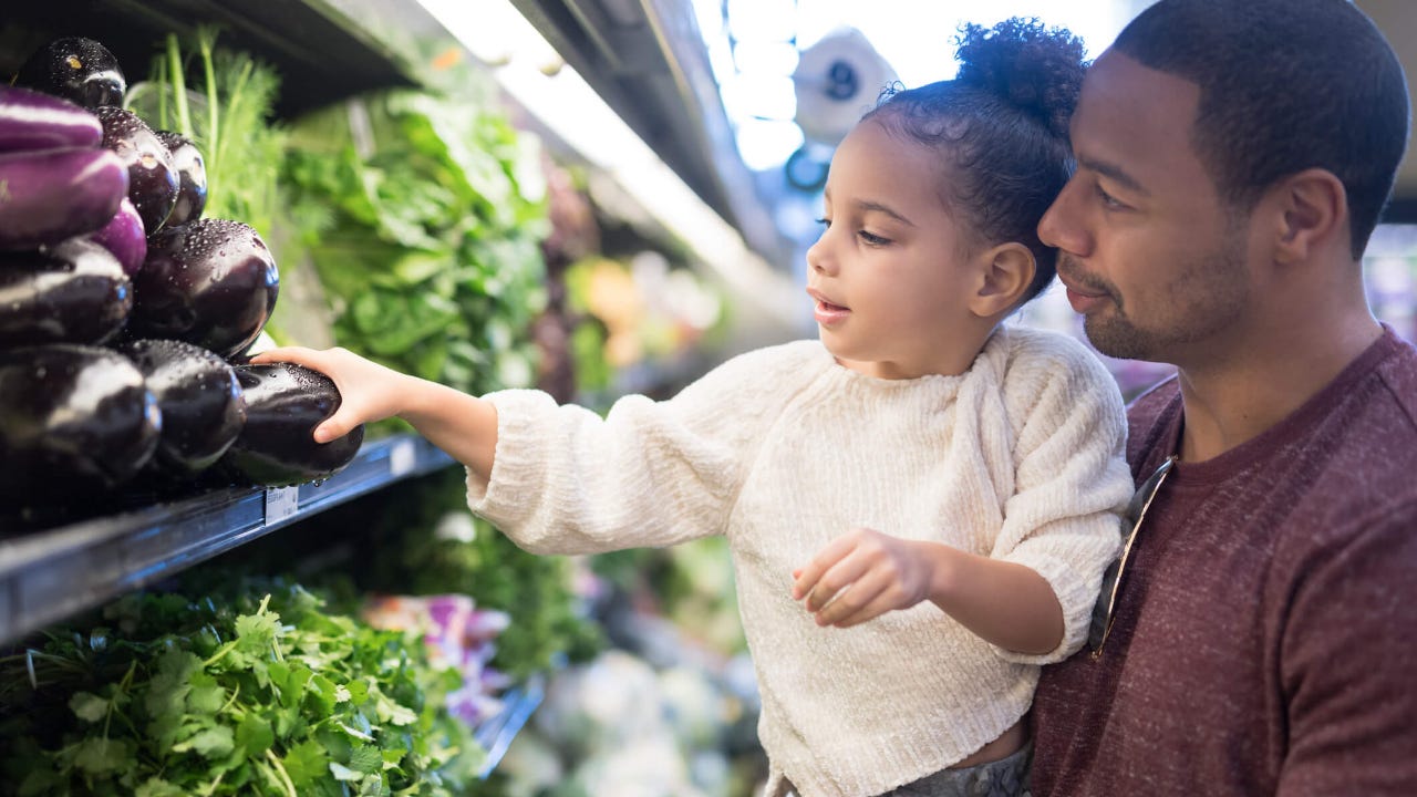 Father holds young daughter up to pick out eggplant at a grocery store