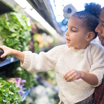 Father holds young daughter up to pick out eggplant at a grocery store