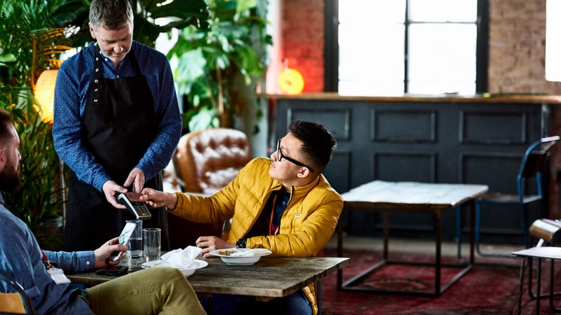 Man paying for lunch at restaurant table with waiter and friend