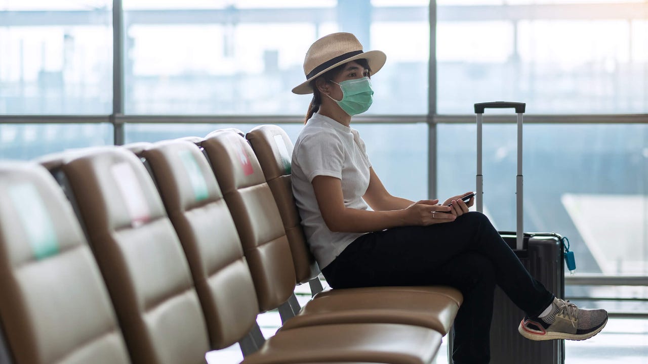 Woman wearing brimmed hat and face mask sits at an airport gate with her luggage as she waits to board