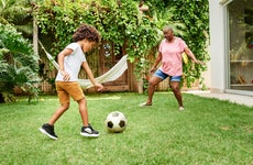A child and their grandparent are kicking a soccer ball in their backyard. There are trees and a hammock in the background.