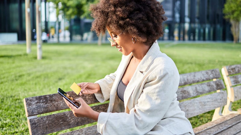 Young woman sitting on a park bench holds her credit card while smiling and using her smartphone