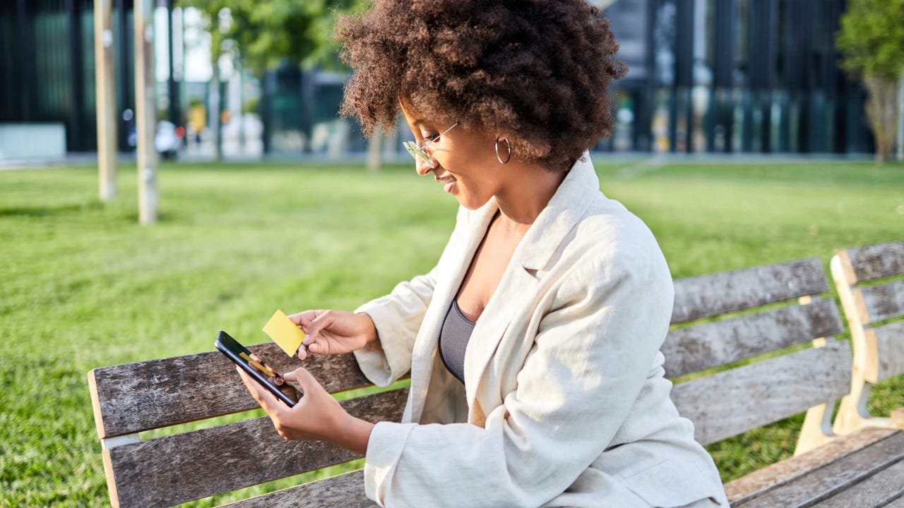 Young woman sitting on a park bench holds her credit card while smiling and using her smartphone
