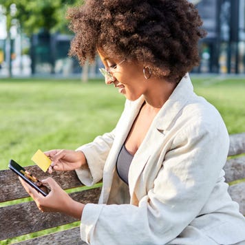 Young woman sitting on a park bench holds her credit card while smiling and using her smartphone
