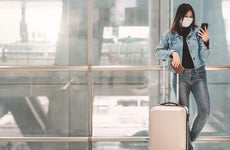 Young woman wearing face mask uses her phone while standing with luggage in airport