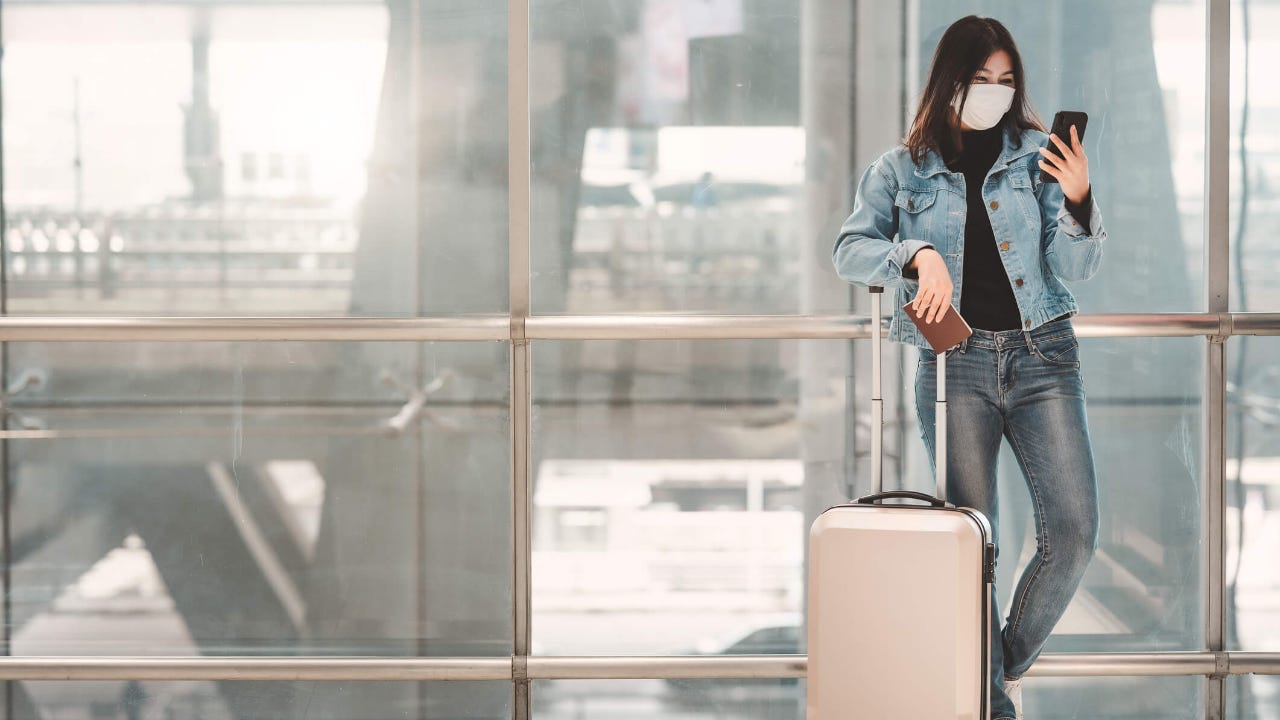 Young woman wearing face mask uses her phone while standing with luggage in airport